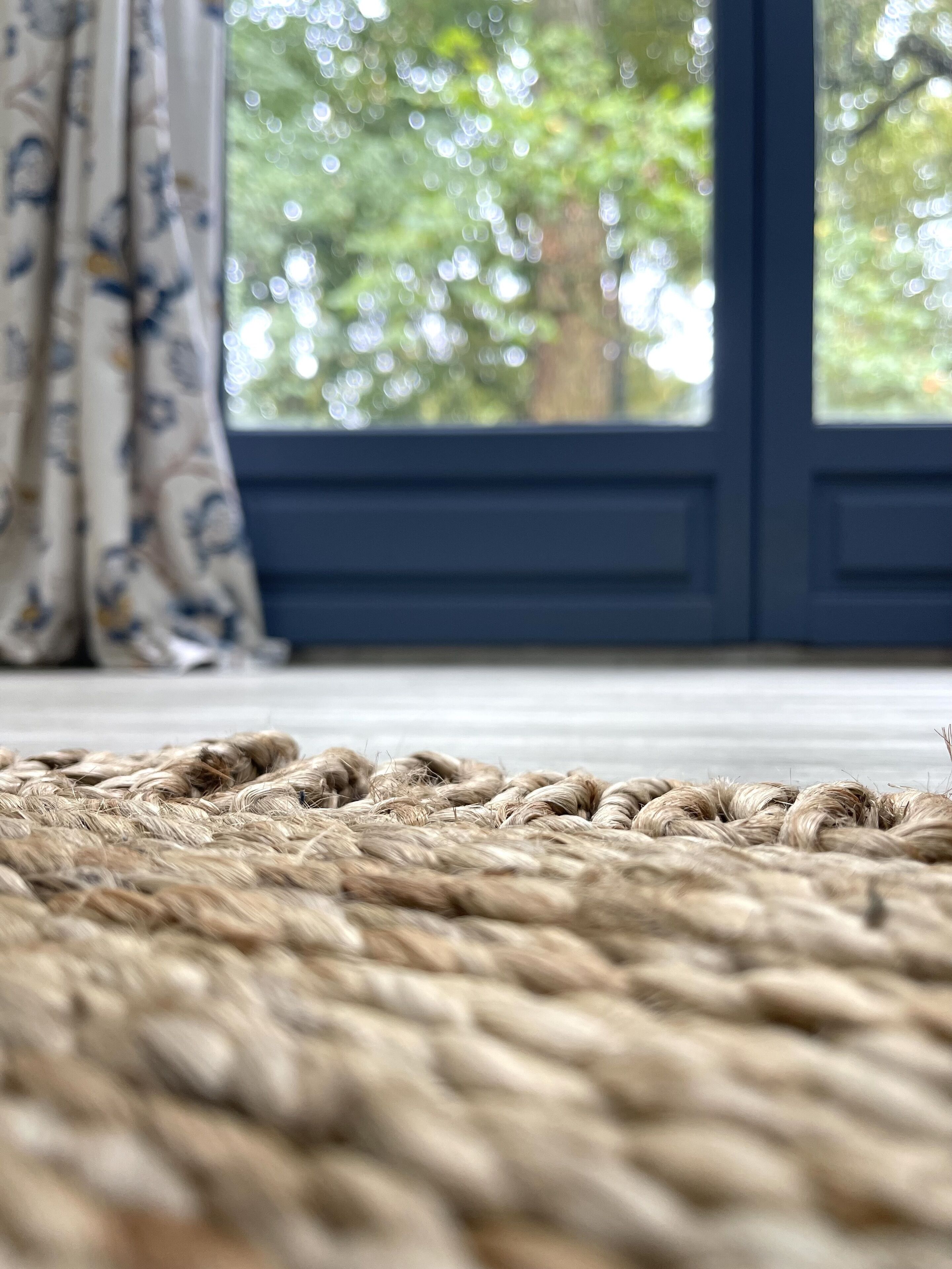 A close-up of a textured rug in a room with a large window, curtains, and a wooden floor.