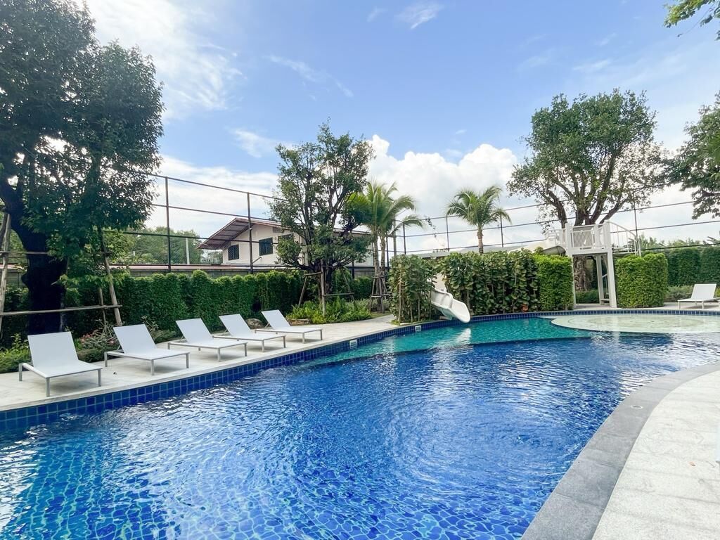 A swimming pool with lounge chairs, surrounded by greenery and a clear sky.