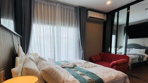A hotel room with a bed, a red armchair, a bedside table with a lamp, and a large window with curtains.