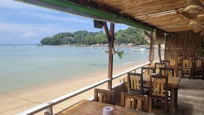 A wooden beachside restaurant with tables and chairs, overlooking a beach and a forested island.