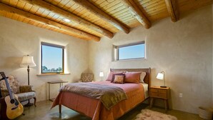 Une chambre avec un plafond en bois, un lit recouvert d’une couverture rose, une guitare, une lampe et une petite table.