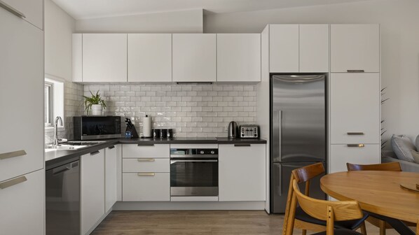 A modern kitchen with white cabinetry, stainless steel appliances, and a wooden dining table. - Remarkable Woodsider - family friendly walk to town (Queenstown)