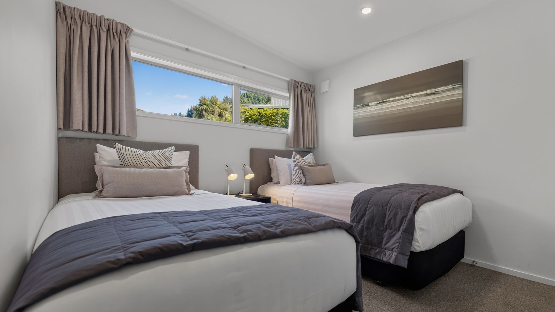A bedroom with two beds, a window with curtains, a bedside table with lamps, and a painting on the wall.
