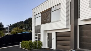 Un bâtiment moderne à deux étages, doté d’un portail de garage, d’un petit jardin à l’avant et offrant une vue sur une zone boisée en arrière-plan.