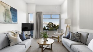 A modern living room with two grey sofas, a coffee table, a TV, a lamp, and a view of a mountain and trees through large windows.