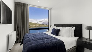 A modern hotel room with a large bed, a view of a lake and mountains, and a window with curtains. - Belle Vue Villa - Stunning Views with Hot Tub (Queenstown)