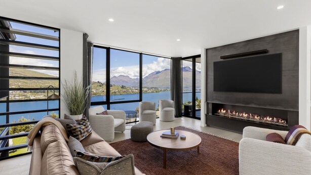 A modern living room with a large TV and fireplace, large windows with a mountain view, and comfortable seating. - Belle Vue Villa - Stunning Views with Hot Tub (Queenstown)