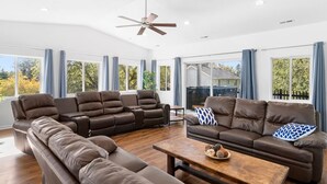 Amplia sala de estar con sofá seccional, sofá más pequeño y una mesa de centro. Cuenta con ventilador de techo y ventanas grandes con cortinas.