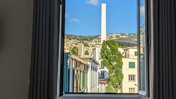 A window view of a cityscape with buildings, a prominent white tower, and a tree in the foreground. - Tanoeiros Studios (Funchal)