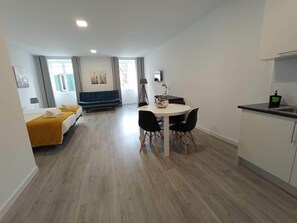 A modern hotel room with a bed, a sofa, a dining table with chairs, a small kitchen area, and a large window with curtains. - Tanoeiros Studios (Funchal)