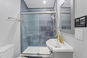 Ein modernes Badezimmer mit einer gläsernen Duschkabine, einem weißen Waschbecken mit goldener Armatur und einem Spiegel darüber.