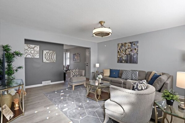 Ein modernes Wohnzimmer mit einem grauen Sofa, einem grauen Sessel und einem Couchtisch. An der Wand hängt ein Gemälde, und eine Deckenleuchte sorgt für Licht.