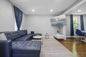 A modern living room with a dark blue sectional sofa, a small round coffee table, and a white side table. There is a large window with curtains, a dining area with blue chairs, and a painting on the wall. - A Dream Home in Chicagoland (Homewood)