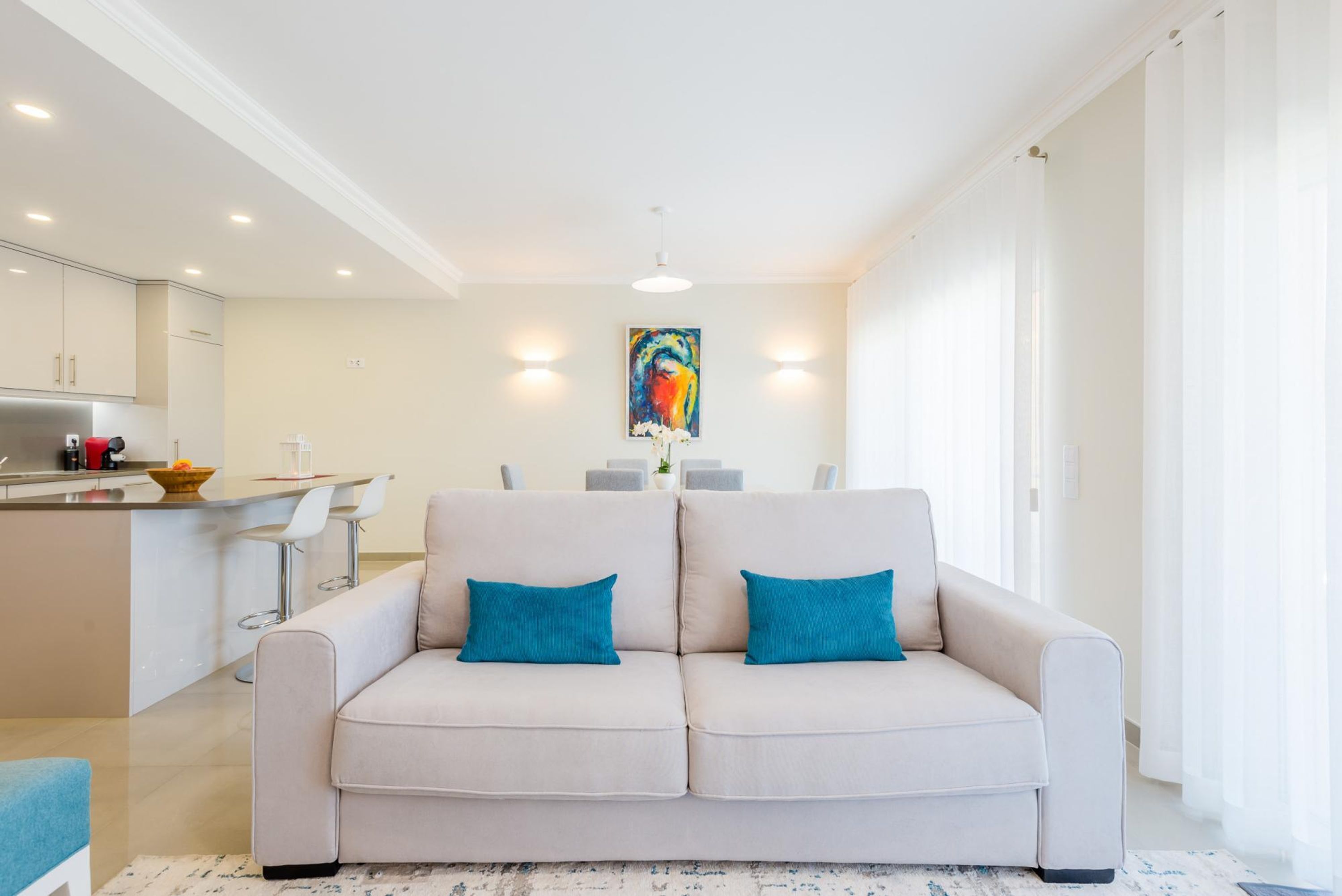 A modern living room with a beige sofa, two teal cushions, a kitchen area with white cabinets, and a dining table with chairs.
