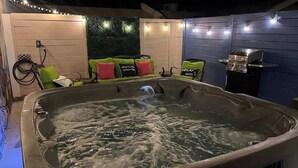 A hot tub with a waterfall feature, surrounded by outdoor seating with green and red cushions, and a barbecue grill.