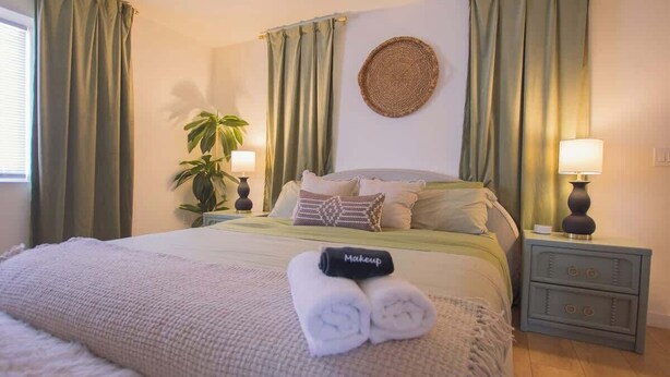 A neatly made bed with a folded towel and a makeup bag, a bedside lamp, a wall-mounted woven item, and a potted plant.