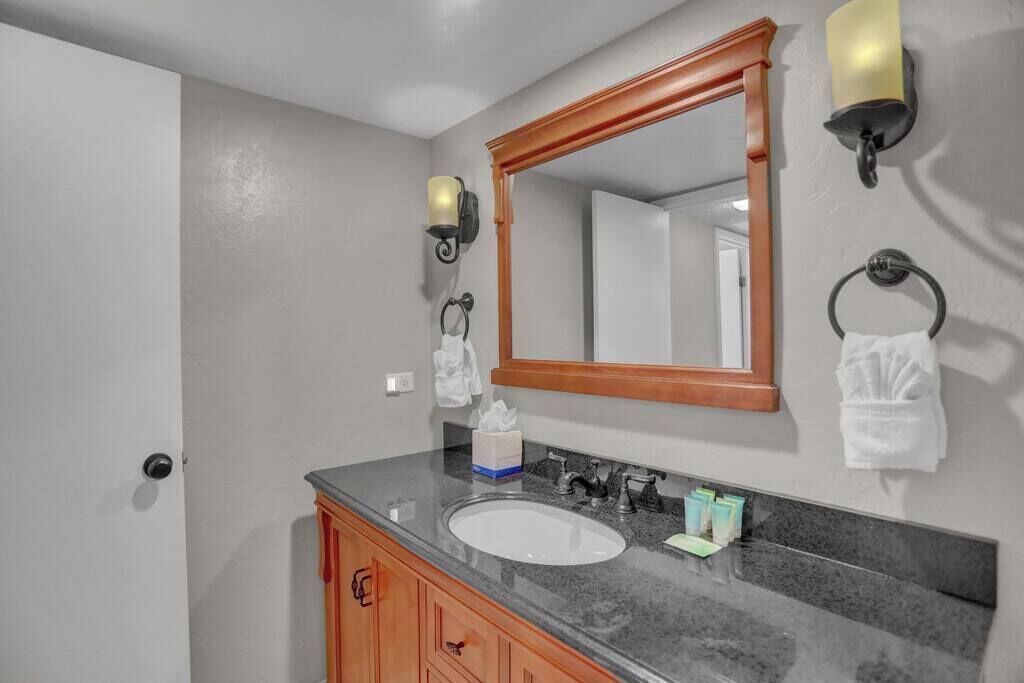 Une salle de bain avec un lavabo blanc, un meuble-lavabo en bois et un miroir avec un cadre en bois. On y trouve deux lampes fixées au mur et un porte-serviettes avec des serviettes.