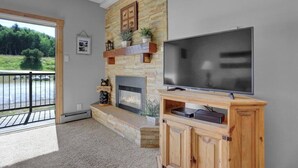 Un salon doté d’une cheminée en pierre, d’un grand téléviseur à écran plat posé sur un meuble en bois, et offrant une vue sur un paysage verdoyant grâce à une porte vitrée.