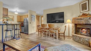 A cozy living room with a fireplace, a dining area with wooden chairs, a kitchen with a refrigerator, and a flat-screen TV mounted on the wall. - Pines Peak Place Summ (Steamboat Springs)
