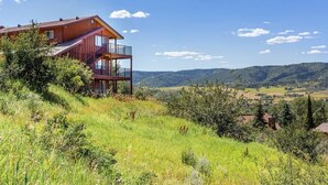 A two-story house with a balcony, surrounded by lush greenery and a scenic view of rolling hills.