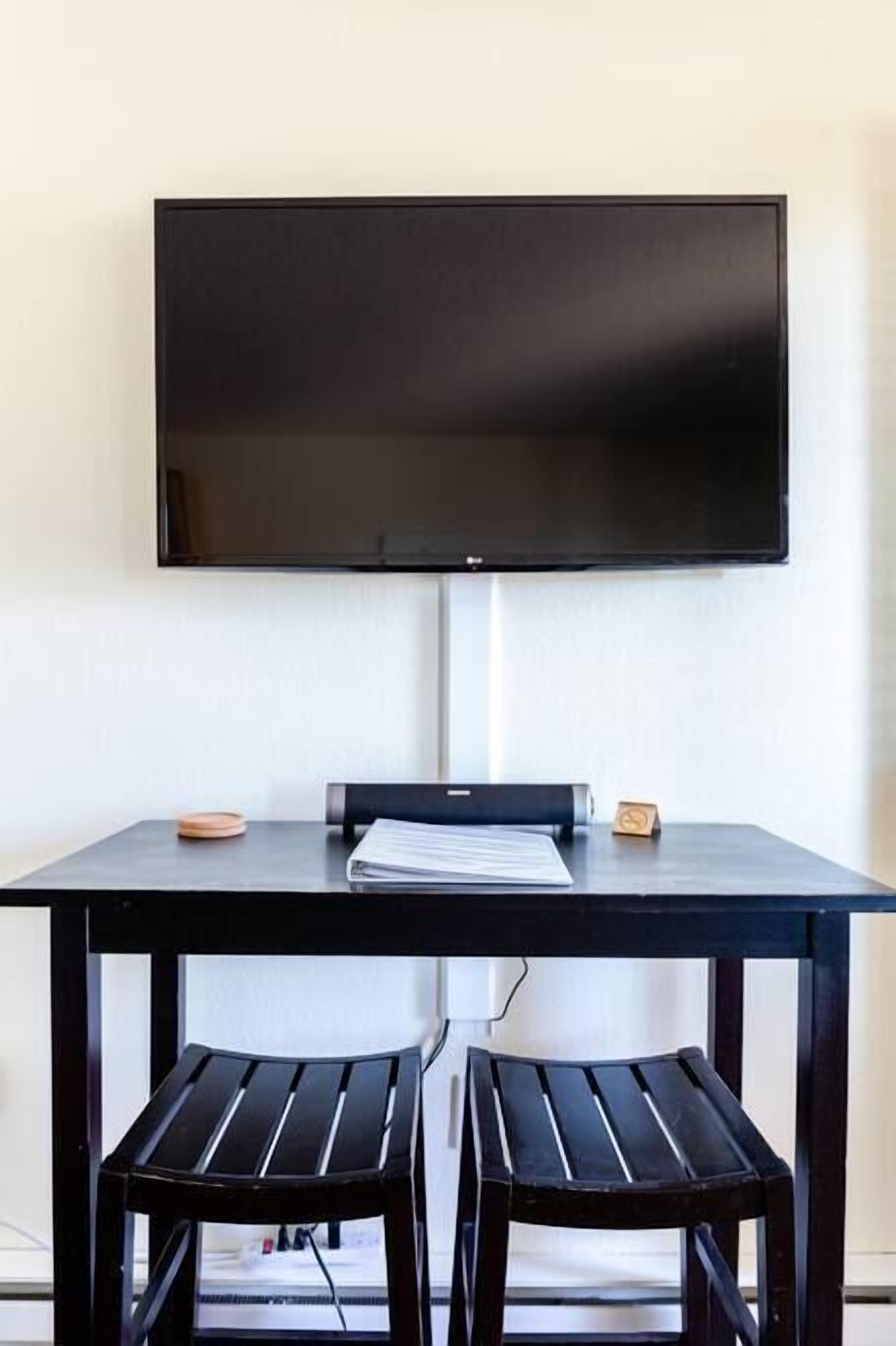 A room with a flat-screen TV mounted on the wall, a black table with two black chairs, and a small wooden block on the table.