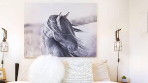 A bedroom with a large horse portrait on the wall, a white bed with a fluffy pillow, and two wall-mounted lamps.