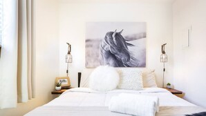 A neatly made bed with a white comforter and a fluffy pillow, a framed picture on the nightstand, and a wall-mounted light fixture.
