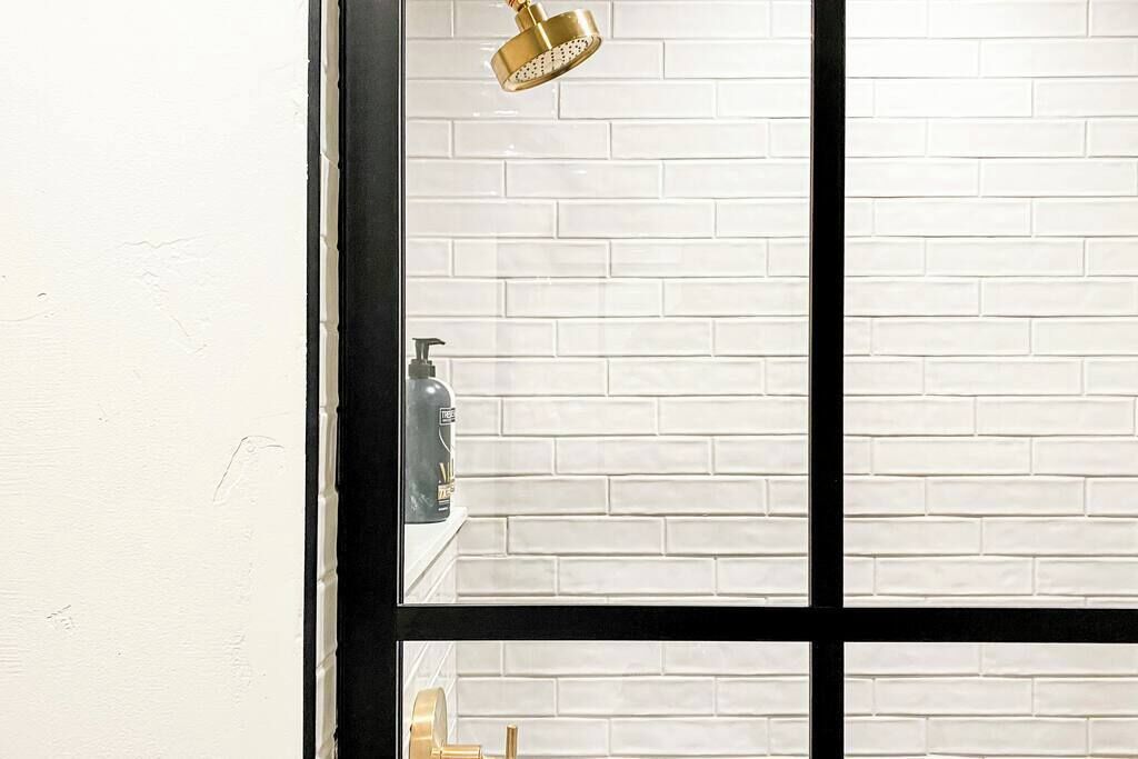 Una ventana con marco negro, pared de ladrillo blanco y cabezal de ducha dorado.