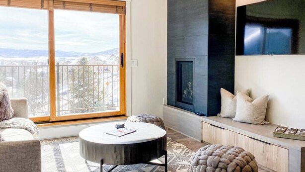 A modern living room with a large window, a round coffee table, a sofa, a fireplace, and a TV mounted on the wall. - Sunset Retreat (Steamboat Springs)