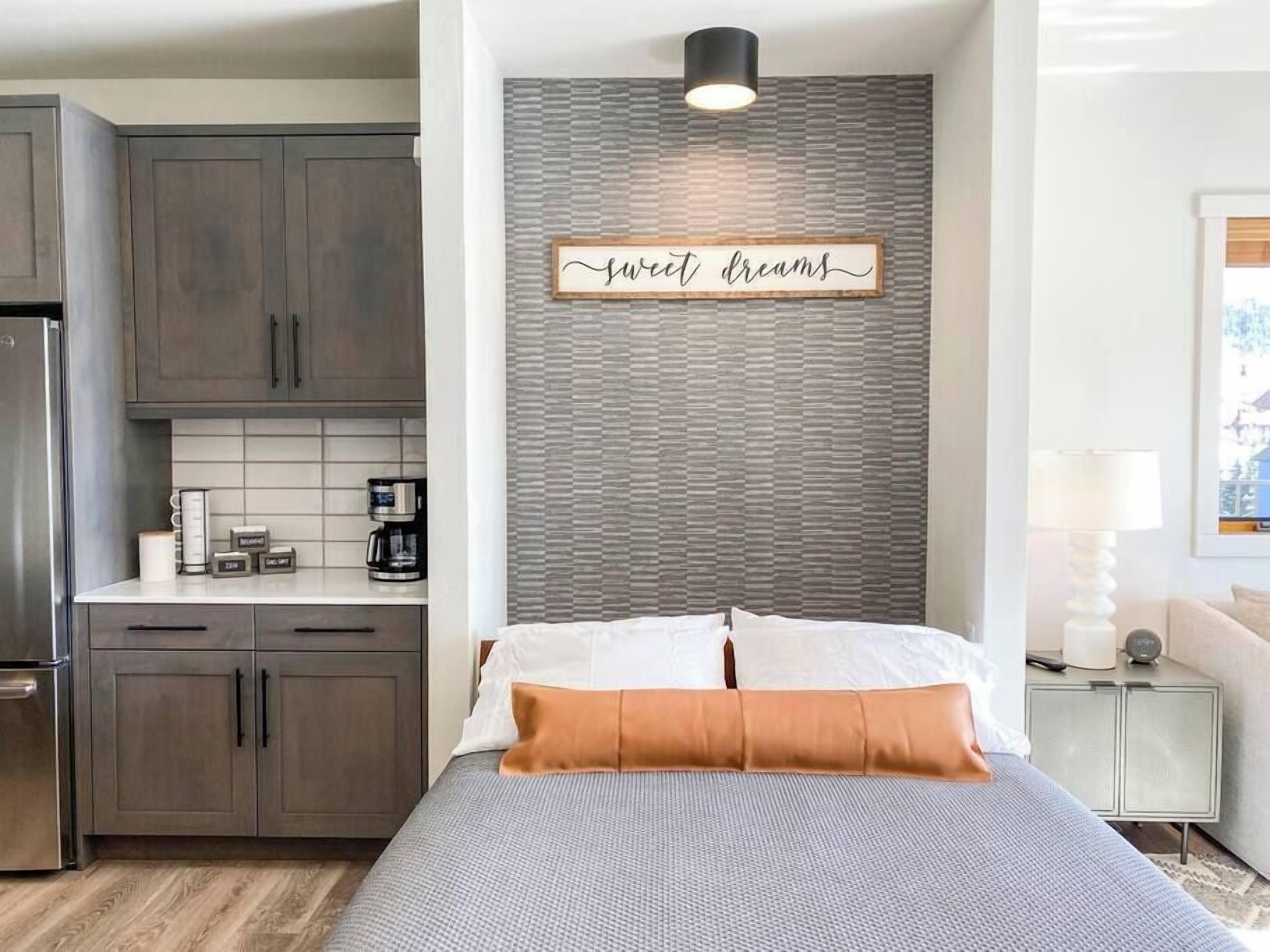 A bedroom with a bed, pillows, a bedside table, a lamp, and a wall-mounted sign.
