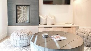 A modern living room with a round coffee table, a flat-screen TV mounted on the wall, a built-in bench with cushions, and a window with blinds. - Sunset Retreat (Steamboat Springs)