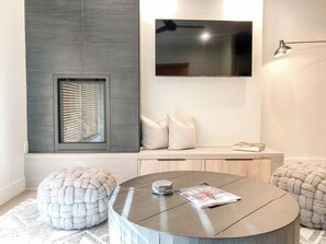 A modern living room with a round coffee table, a flat-screen TV mounted on the wall, a built-in bench with cushions, and a window with blinds.