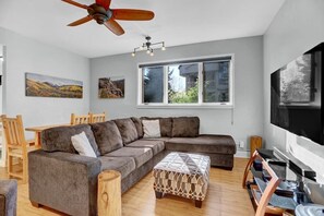 Ein modernes Wohnzimmer mit einem Ecksofa, Holzböden, einem Deckenventilator, einem großen Fenster und einem Flachbildfernseher.