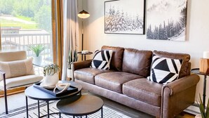 Een woonkamer met een bruine leren bank, een ronde salontafel en een beige fauteuil. Twee ingelijste foto's hangen aan de muur en er staat een vloerlamp.
