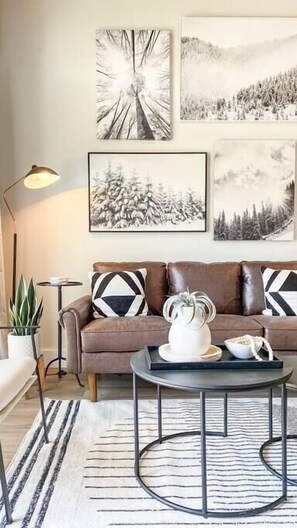 A living room with a brown sofa, two black and white pillows, a round coffee table with a white pitcher, a floor lamp, and a potted plant.
