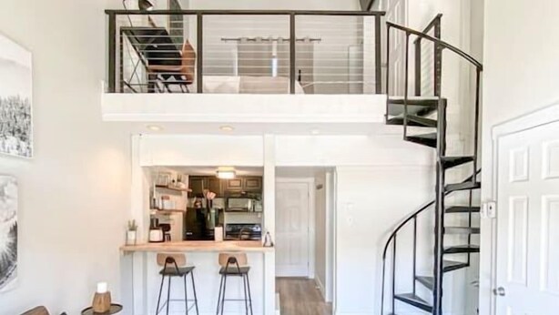 Een ruime woonkamer met een hoge plafond, een trap en een keuken zichtbaar door een open deur.