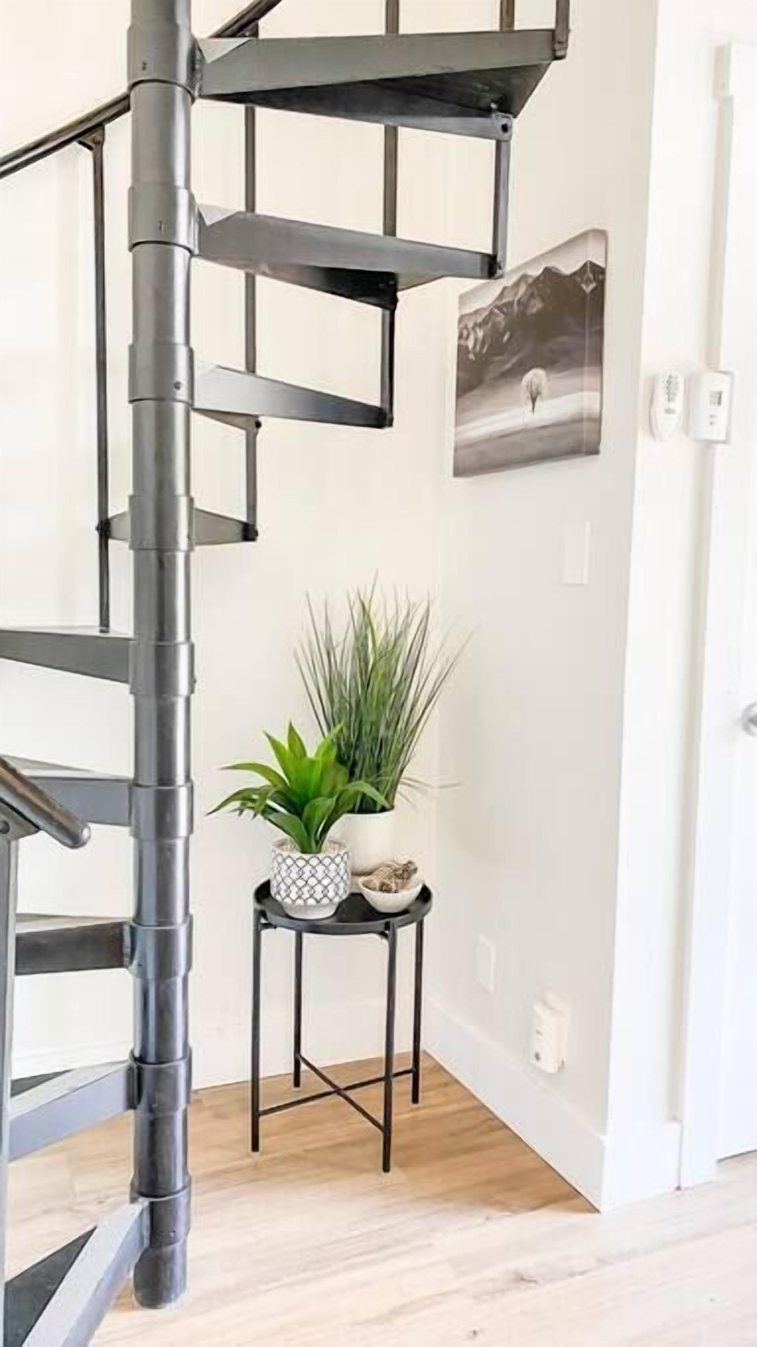 Una escalera moderna con barandilla de metal negro, una mesita pequeña con una planta y objetos decorativos, y un cuadro en la pared.