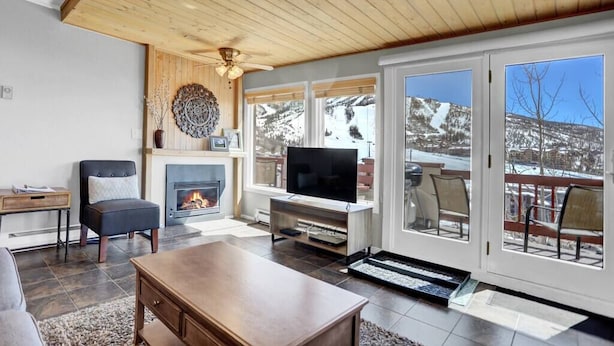 Un salon chaleureux avec une cheminée, un téléviseur et une vue sur des montagnes enneigées, visible à travers de grandes fenêtres.