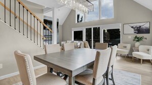 A spacious dining area with a large table and multiple chairs, a staircase with wooden railings, and large windows offering views of the outdoors.