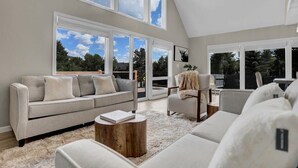 A spacious living room with a grey sofa, a round wooden coffee table, and a fluffy rug. Large windows provide a view of the outdoors.