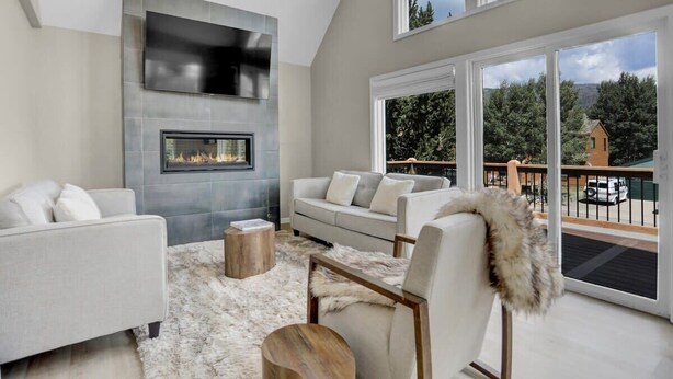 A modern living room with a fireplace, a sofa, a chair, and a view of a deck and trees.