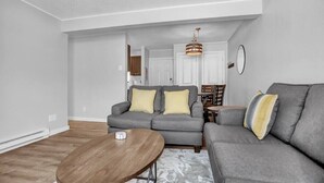 Ein Wohnzimmer mit grauem Sofa, einem Holztisch und einem Essbereich im Hintergrund.