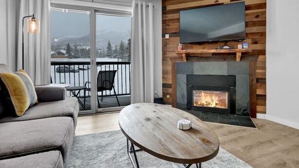 Ein Wohnzimmer mit einer Couch, einem Couchtisch, einem Kamin, einem an einer Holzwand montierten Fernseher und einem Balkon mit Blick auf schneebedeckte Berge.