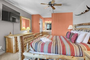 A bedroom with a wooden bed frame, a ceiling fan, a TV, a vanity with a mirror, and a bathroom door visible in the background.