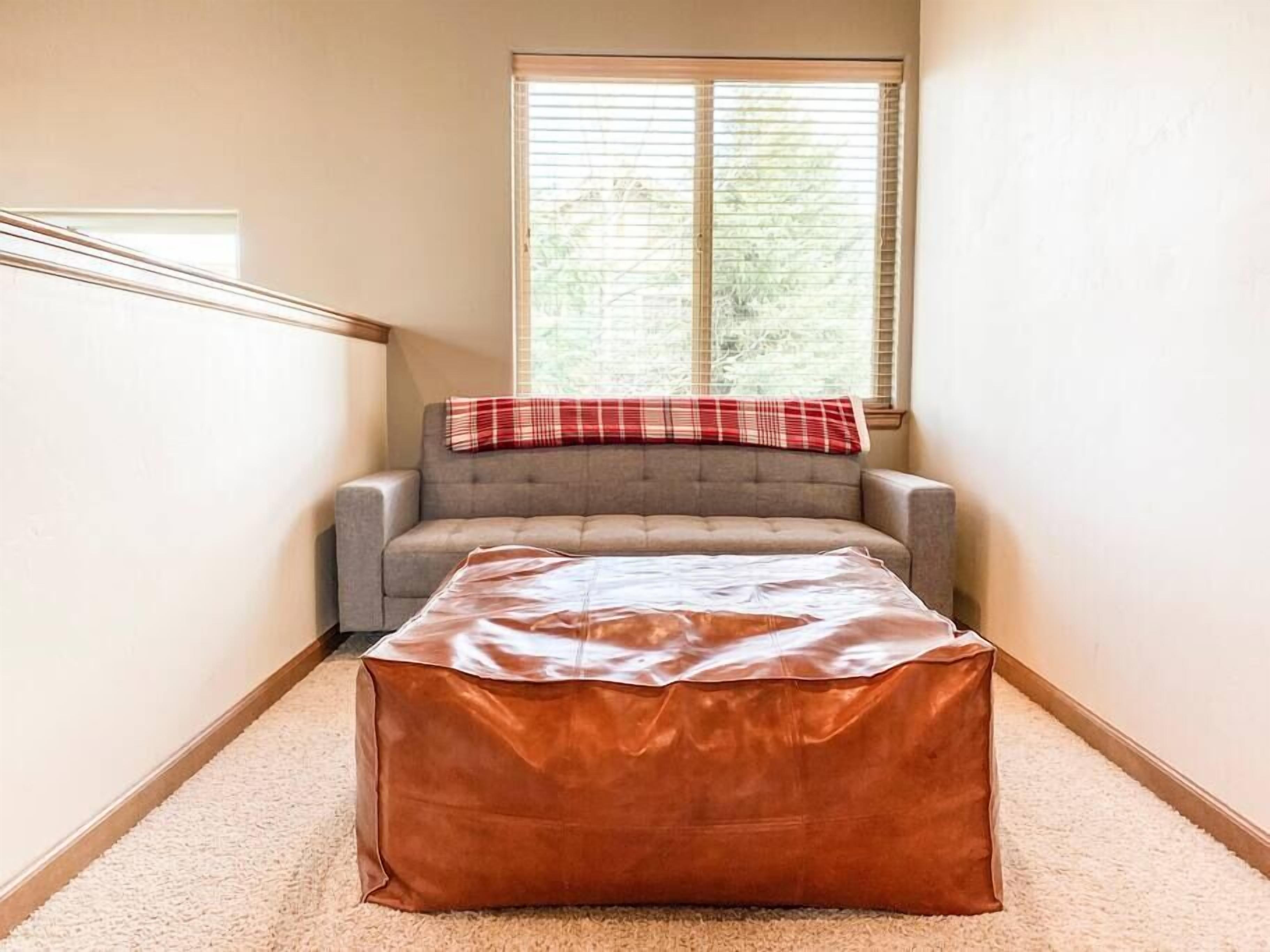 Ein Eckzimmer mit grauem Sofa, einer rot-weißen Decke im Karomuster und einem braunen Ledersitzhocker.