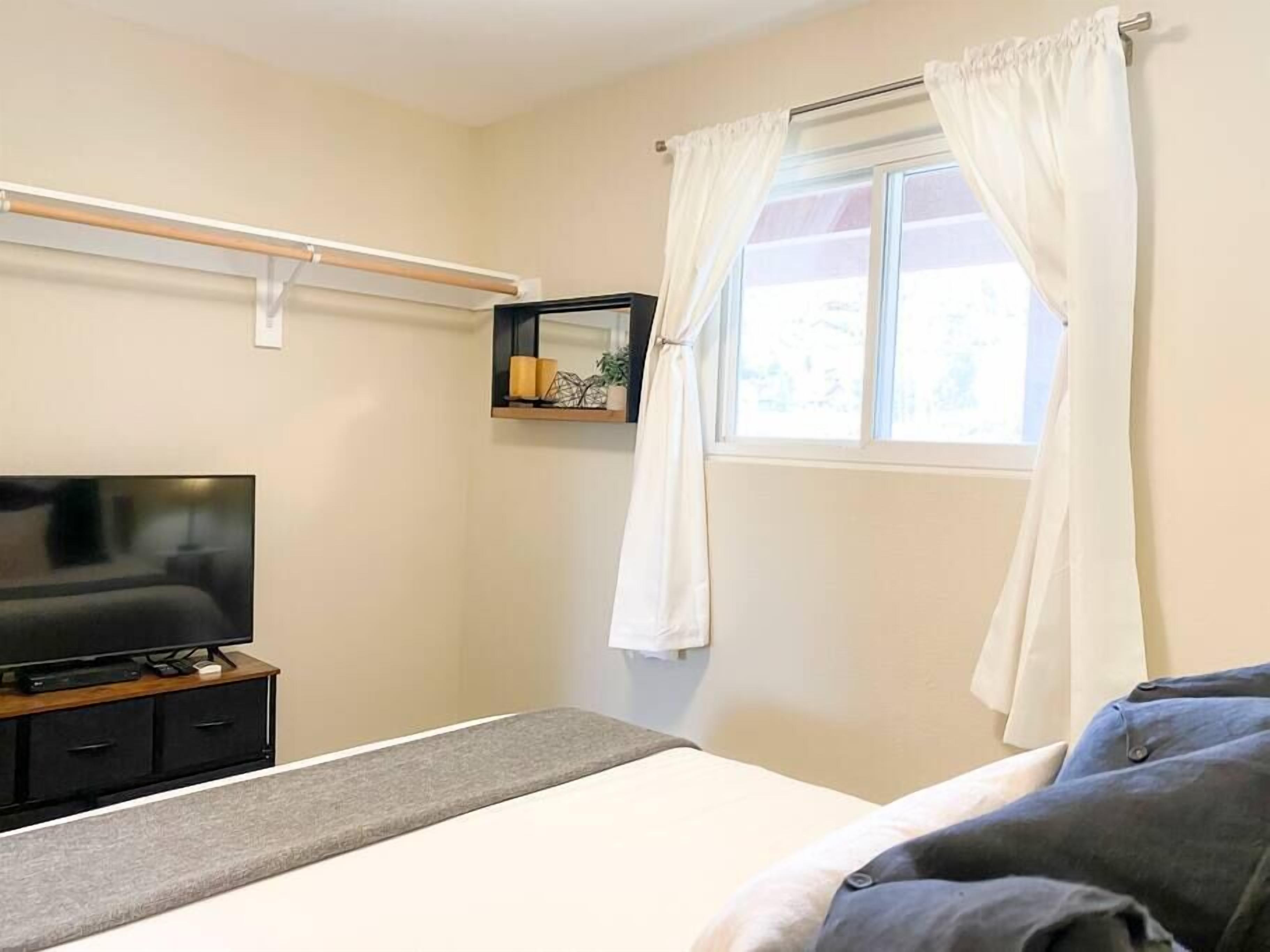 Une chambre à coucher avec un lit, un téléviseur posé sur un meuble en bois, une fenêtre avec des rideaux blancs et une petite étagère ornée d’objets décoratifs.