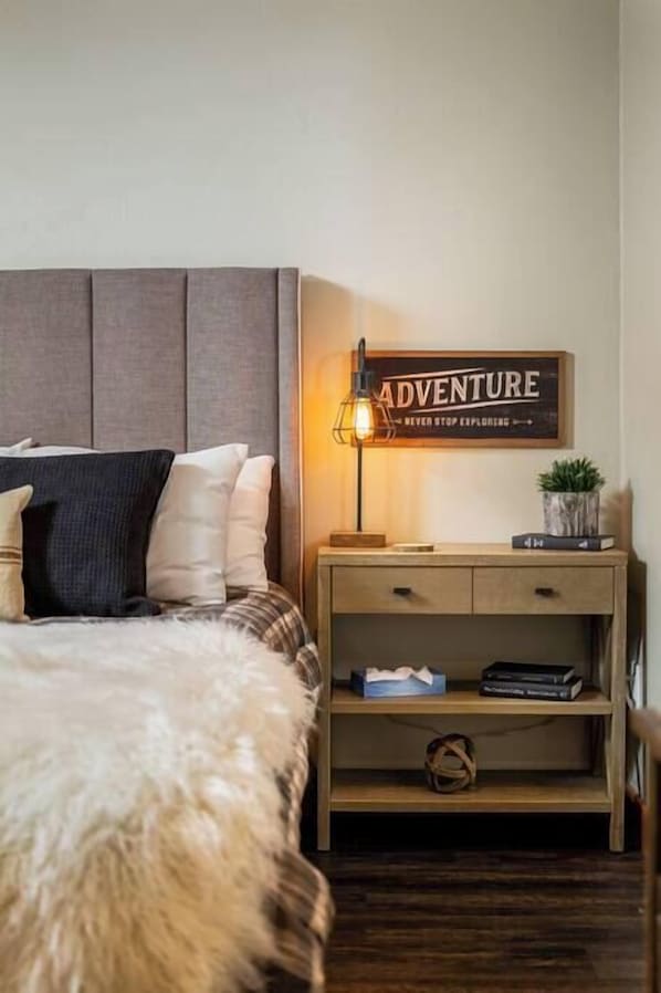 Un dormitorio ordenado con cama, mesita de noche, lámpara y un cartel que dice 'ADVENTURE'.