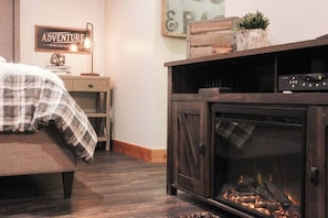 A cozy room with a bed, a wooden cabinet with a fireplace, a small table with a lamp, and a wall sign. - Hillside Haven (Steamboat Springs)