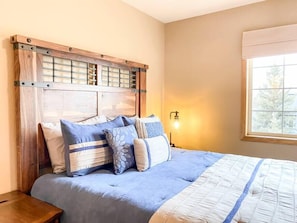 Une chambre à coucher avec une tête de lit en bois, un lit avec une literie bleue, une table de chevet et une fenêtre donnant sur des arbres.