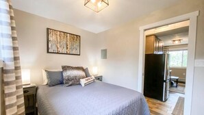 Un dormitorio con cama, mesita de noche, lámpara, un cuadro en la pared y vista a la cocina por una puerta abierta.